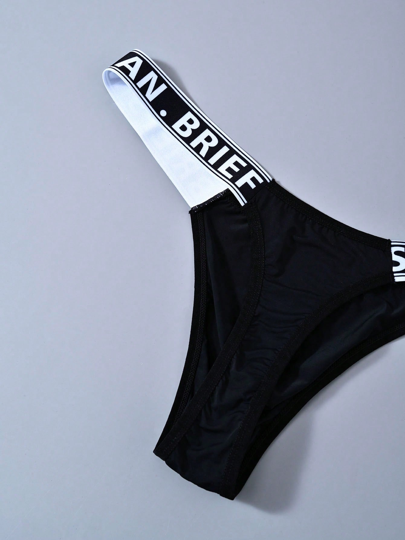 Men's solid color thong underwear with a letter waistband, textured pattern, tape detail, and mid-rise fit