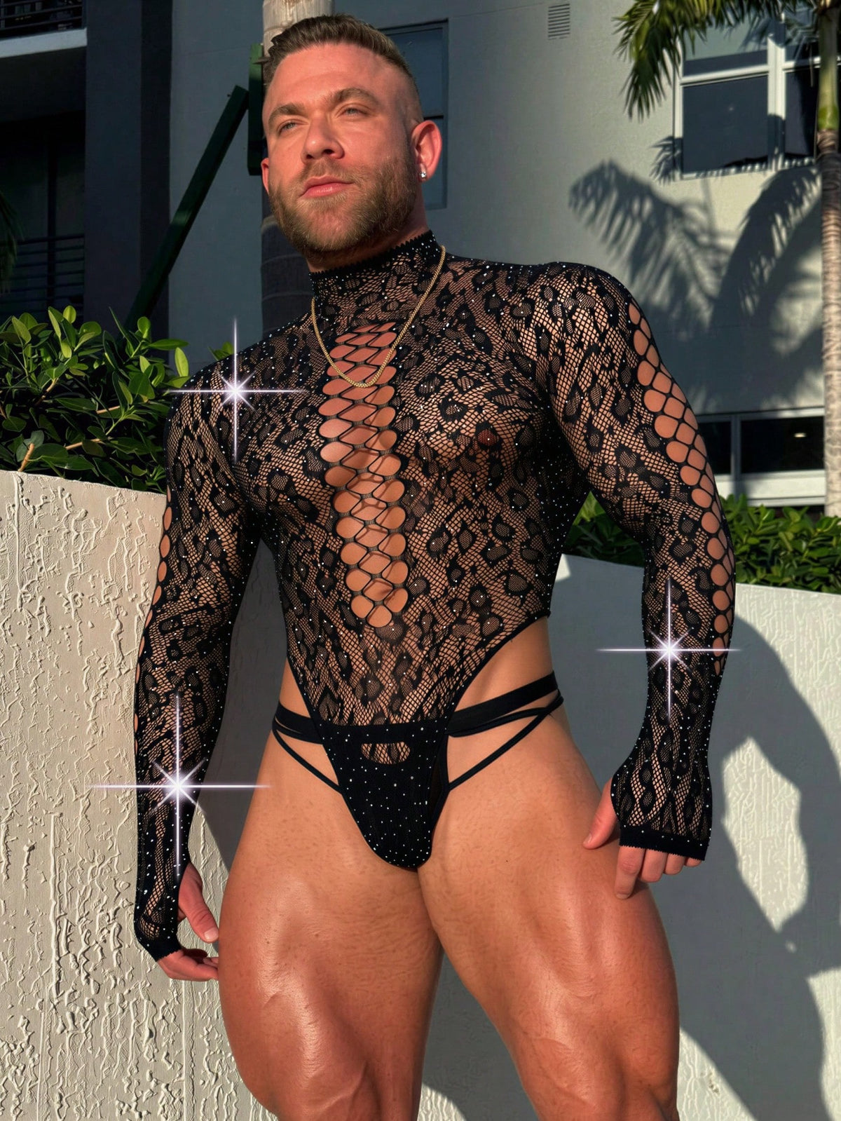 A man wearing a black hollow out rhinestone long jumpsuit featuring a stand collar and sheer knitted fabric.
